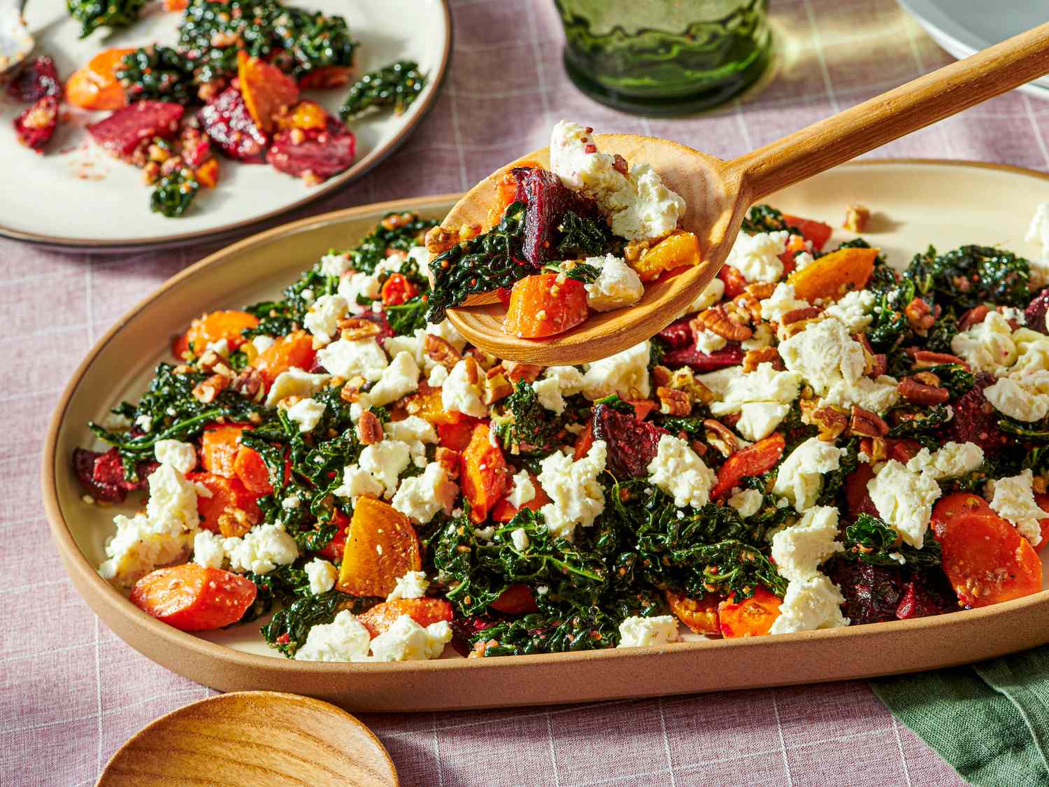 A platter containing kale salad with roasted root vegetables and crumbled cheese served with a wooden spoon