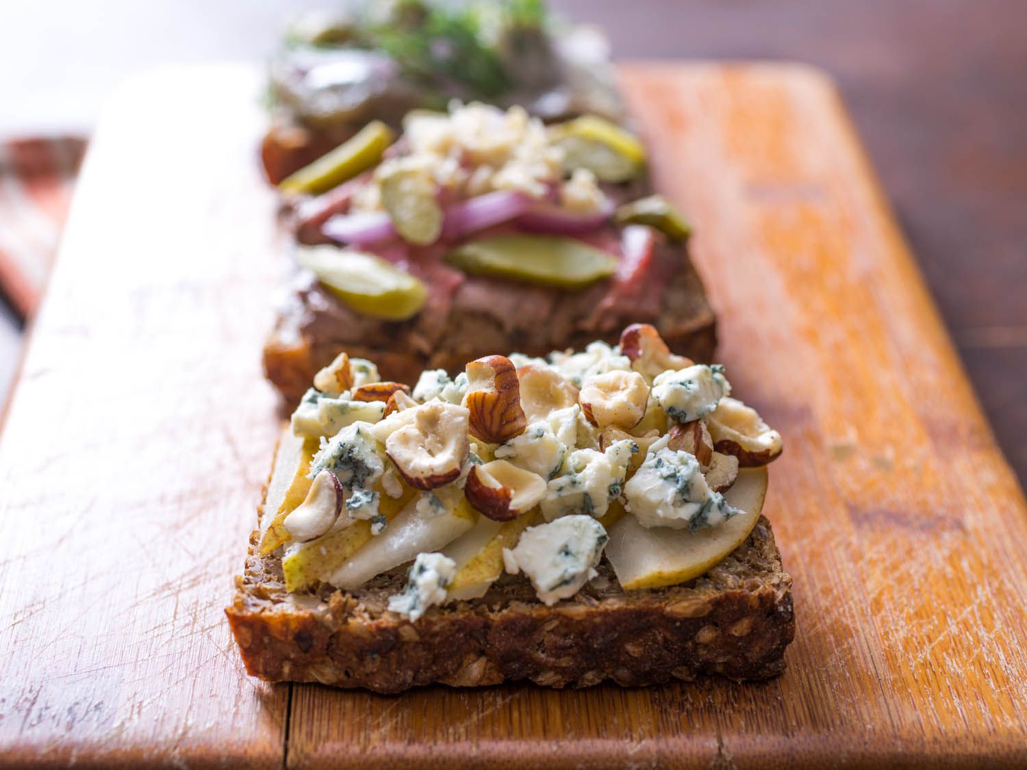 Three different Smørrebrød on a wood cutting board, each with different toppings. The one in the foreground has pear slices, blue cheese crumbles, and chopped hazelnuts.