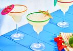 Three margaritas on a blue tile surface, each garnished with small paper umbrellas, with a decorated napkin and lime nearby