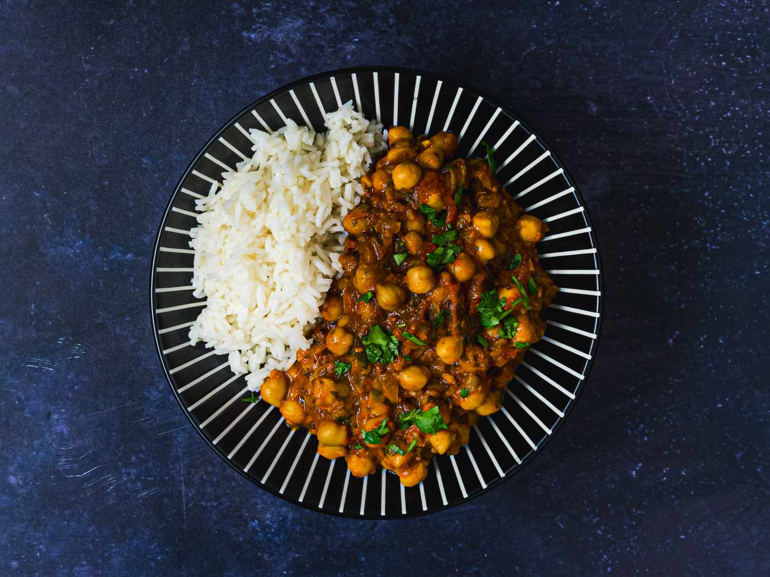 Channa masala and white rice on a blue plate decorated with white lines. The plate is on a dark blue surface.
