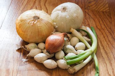 A pile of different types of whole, unpeeled onions on a cutting board. 