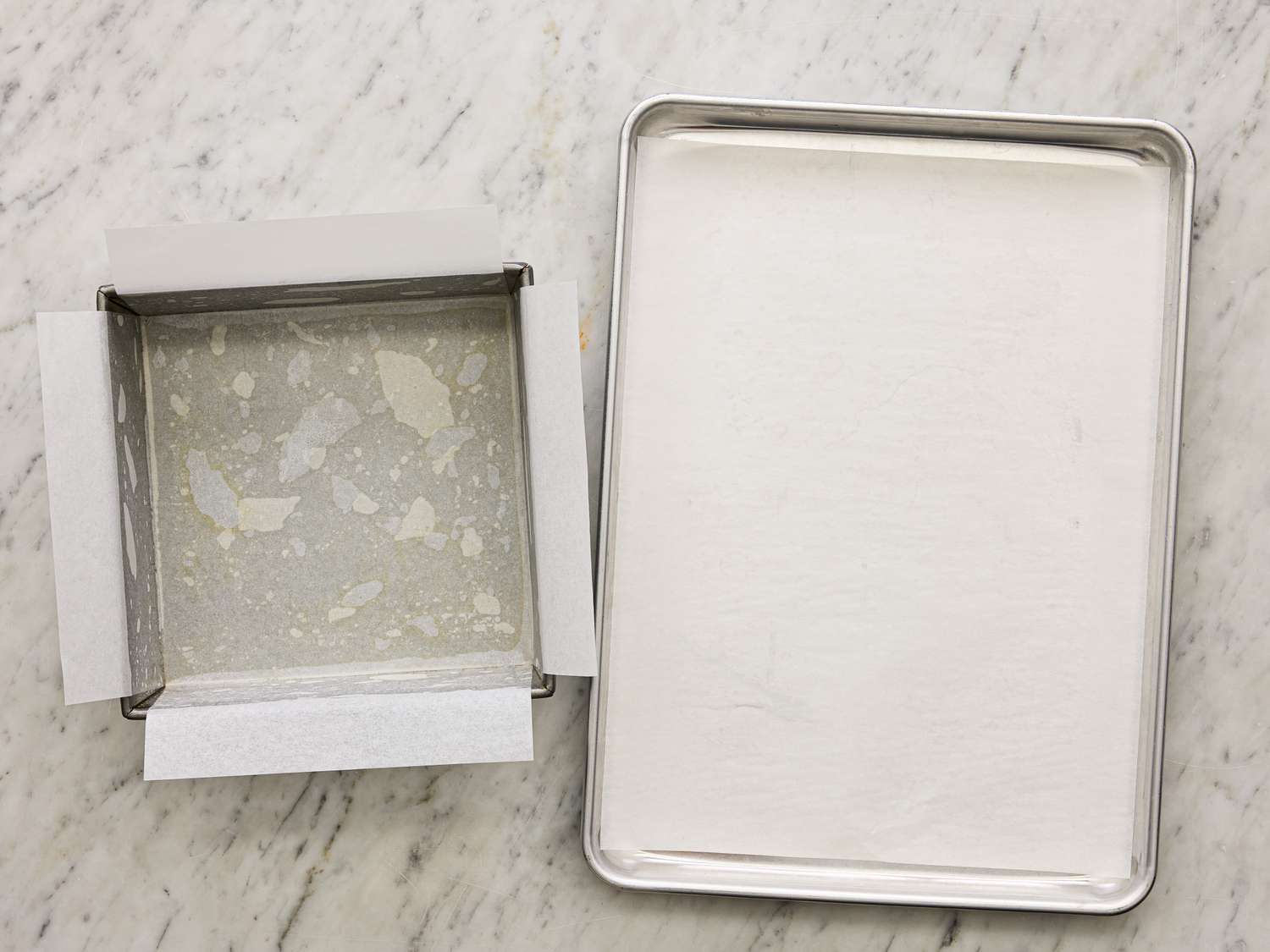 A parchmentlined square baking pan next to a parchmentlined baking sheet on a marble surface