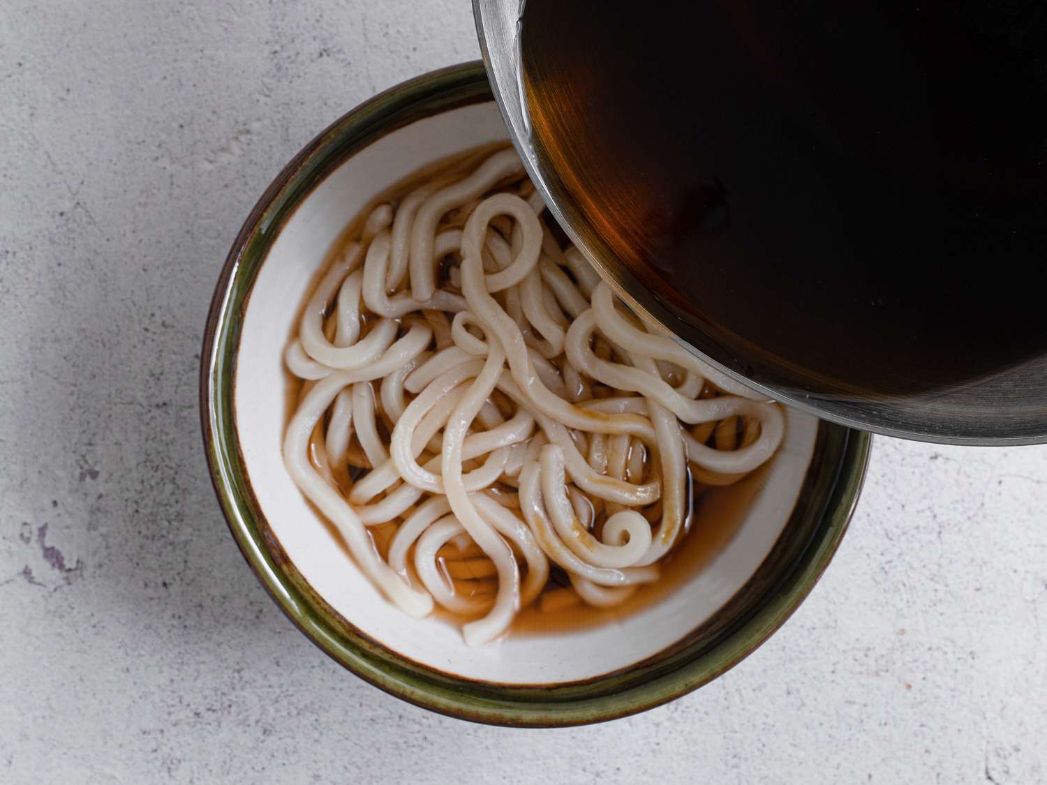 Cooked udon noodles in a ceramic bowl with vegan broth being poured over them.