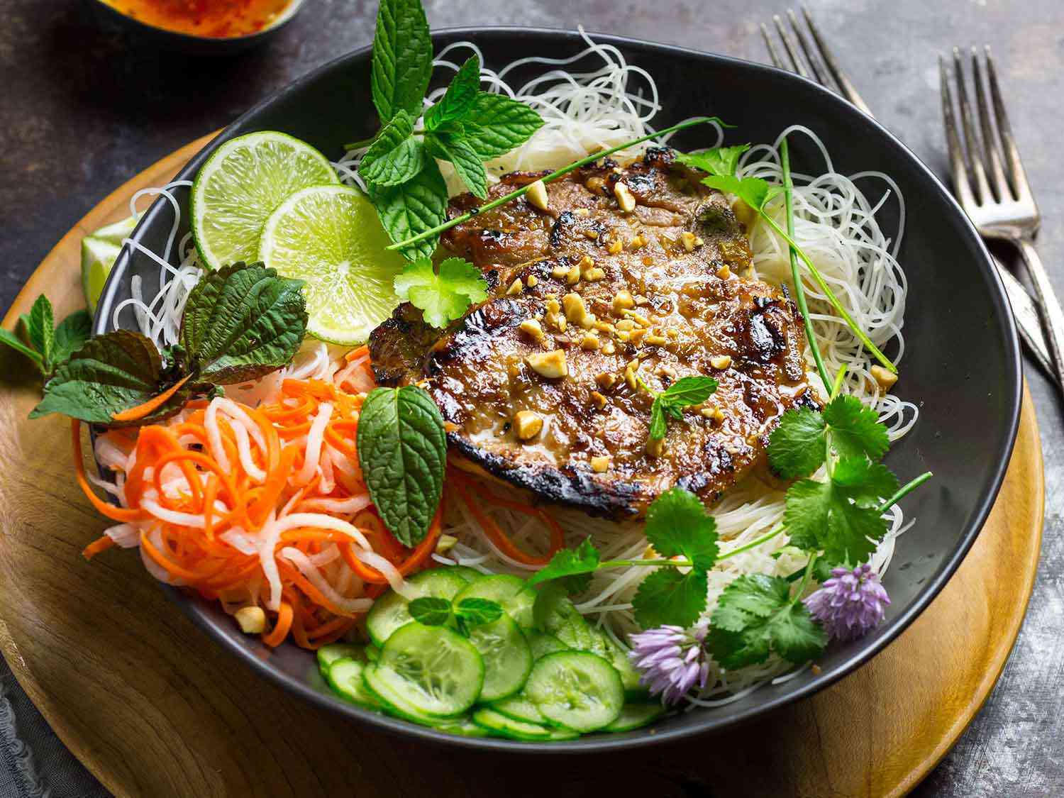 A close-up of a bowl of the vermicelli. A grilled pork chops sits atop the noodles, garnished with crushed peanuts, and pickled and fresh vegetables sit alongside. Cilantro, perilla/shiso, and mint sprigs are scattered over the bowl, along with a few chive blossoms and lime slices.