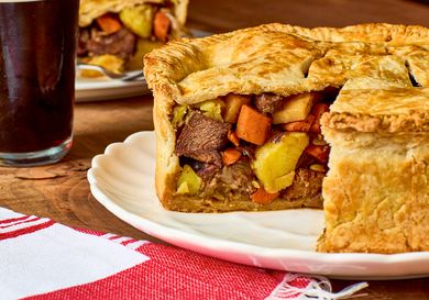 A meat and vegetable potpie with a sliced section showing the filling served on a white plate with a drink in the background