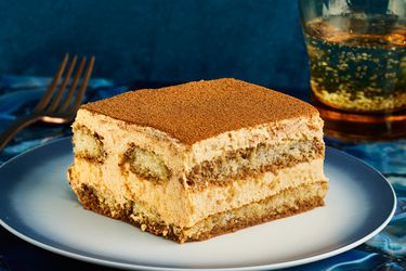 Slice of PSL Tiramisu on a stylish blue and white plate, with a blue tablecloth, sparkling water, and a gold fork