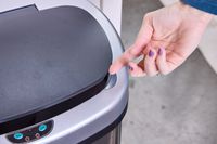 A person lifts the lid on the top of the iTouchless 13-Gallon Oval Stainless Steel Sensor Trash Can