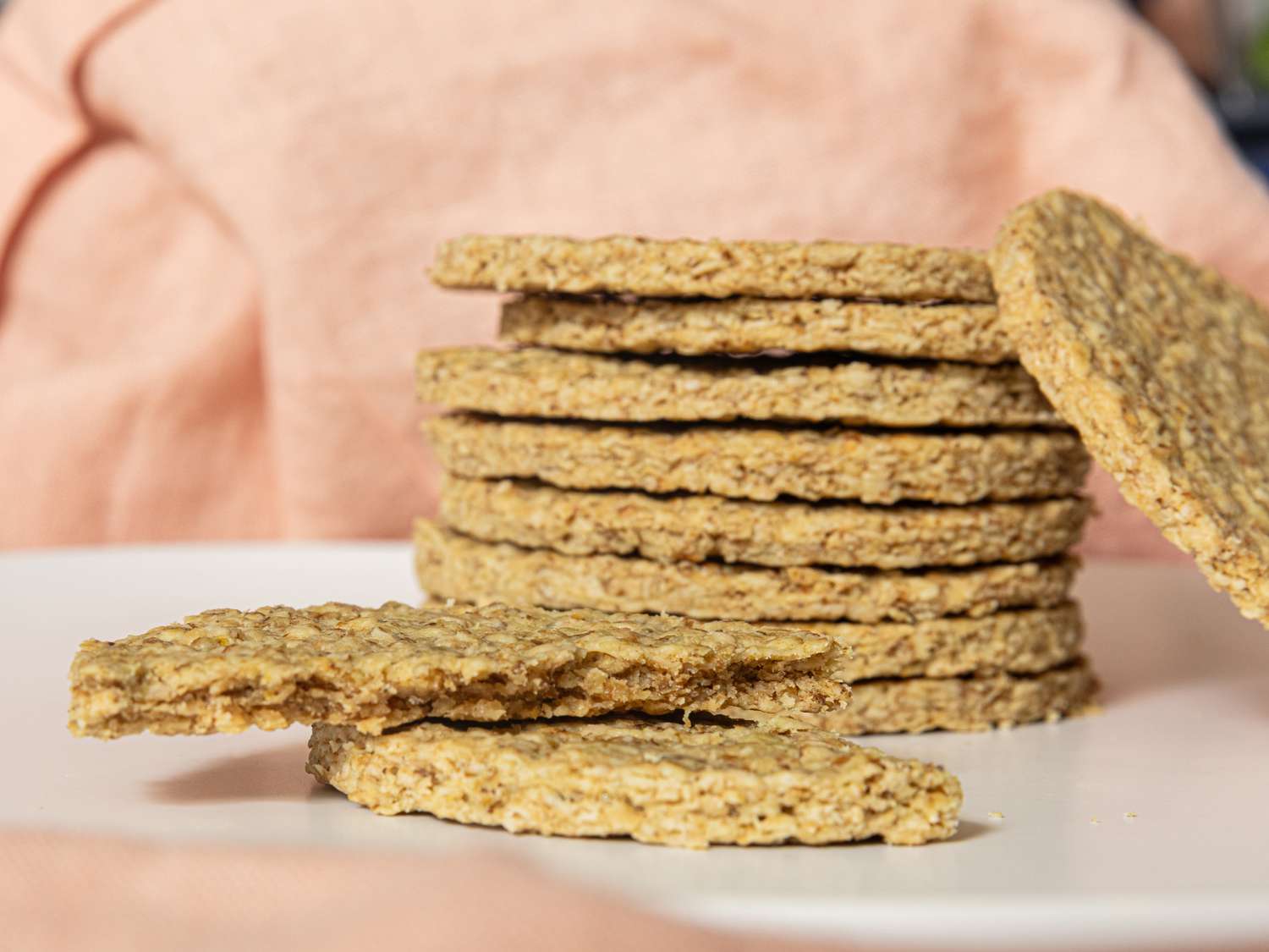 Side view of stack of oatcakes on a white plate, with one oatcake ripped in half. Pink fabric in the backgrounf 