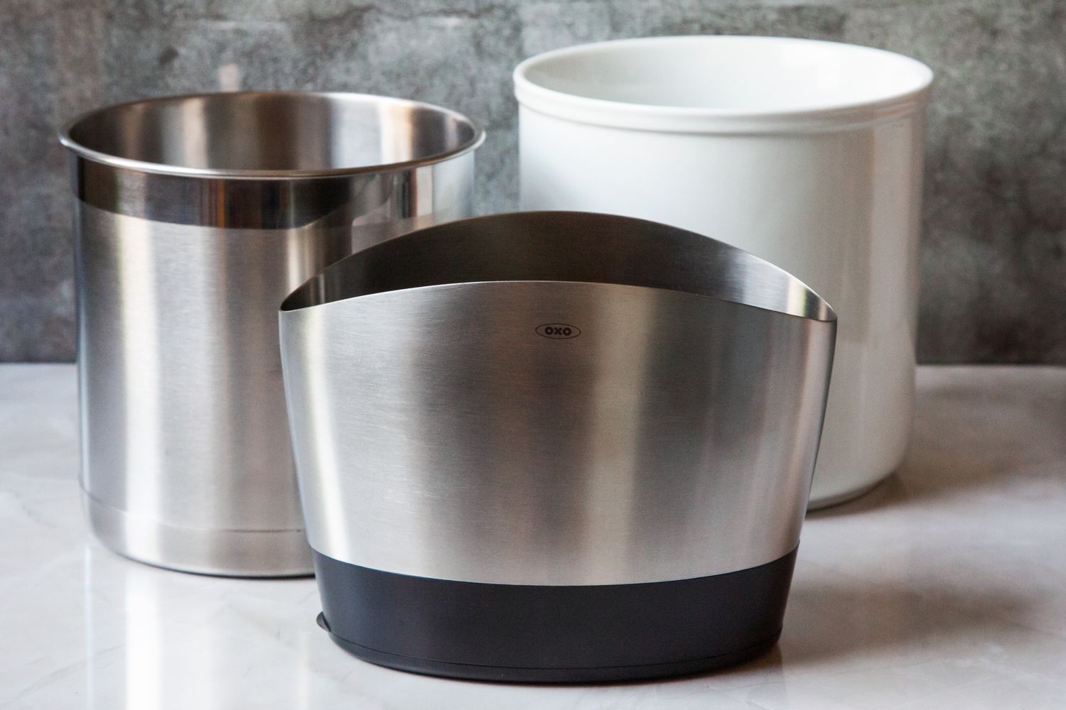 Three utensil crocks (two stainless steel and one ceramic) on a marble kitchen countertop