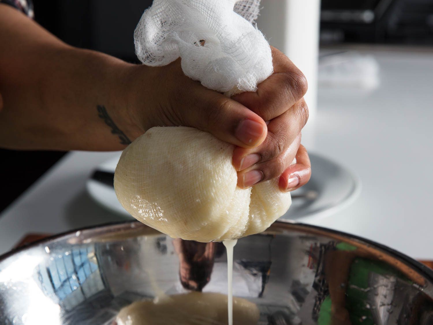 A hand holding potato shreds wrapped in cheesecloth, squeezing out liquid into a metal bowl