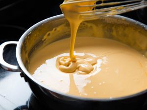 Thick cheese sauce dribbling off of a whisk into a saucepan full of more cheese sauce.