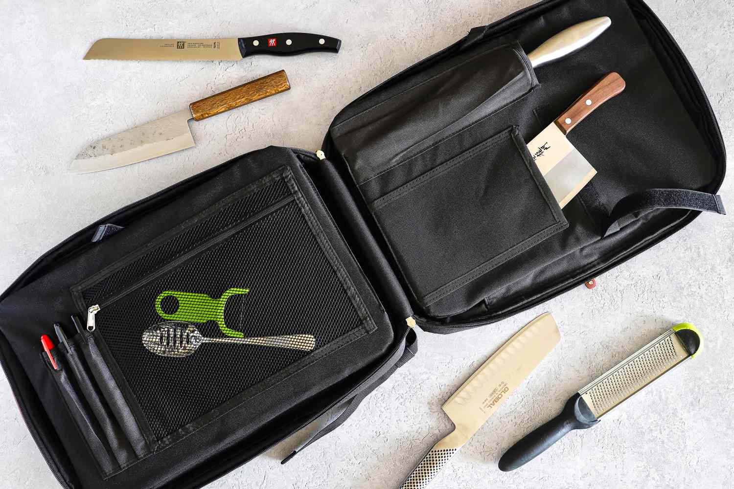A knife and utensil carrying case with kitchen tools and knives laying nearby on the surface