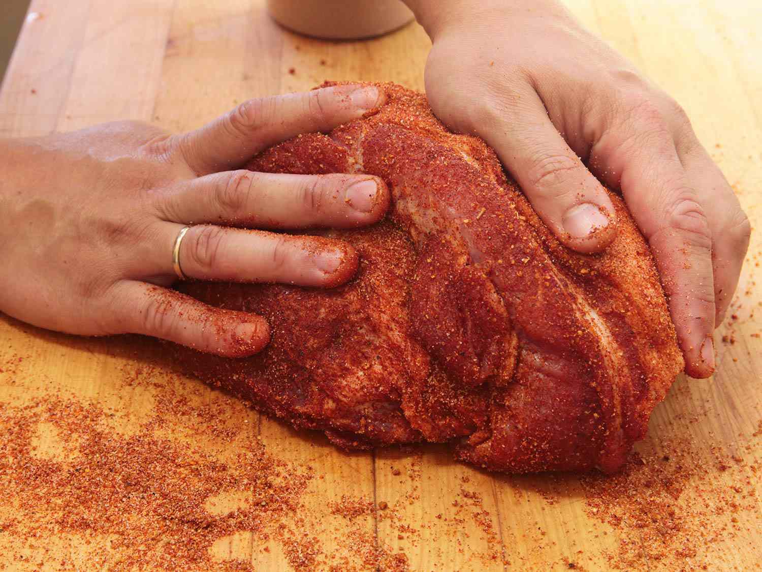 Two hands rubbing seasoning into pork shoulder.