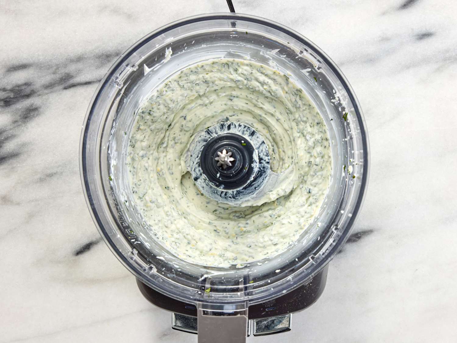 Processing herbs and yogurt in a food processor on a white marble surface 