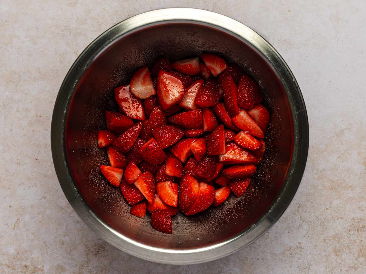 Trimmed and quartered strawberries mixed with sugar inside of a metal mixing bowl.