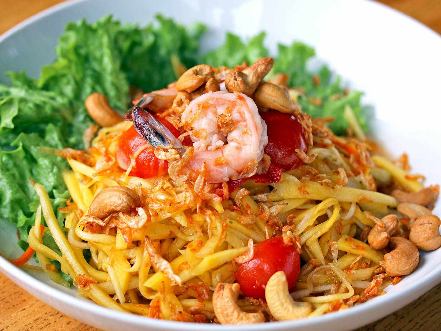 A plate of green-mango salad (tam muk muang) with shredded green mango, cashews, shrimp, tomatoes, and lettuce leaves in the background.