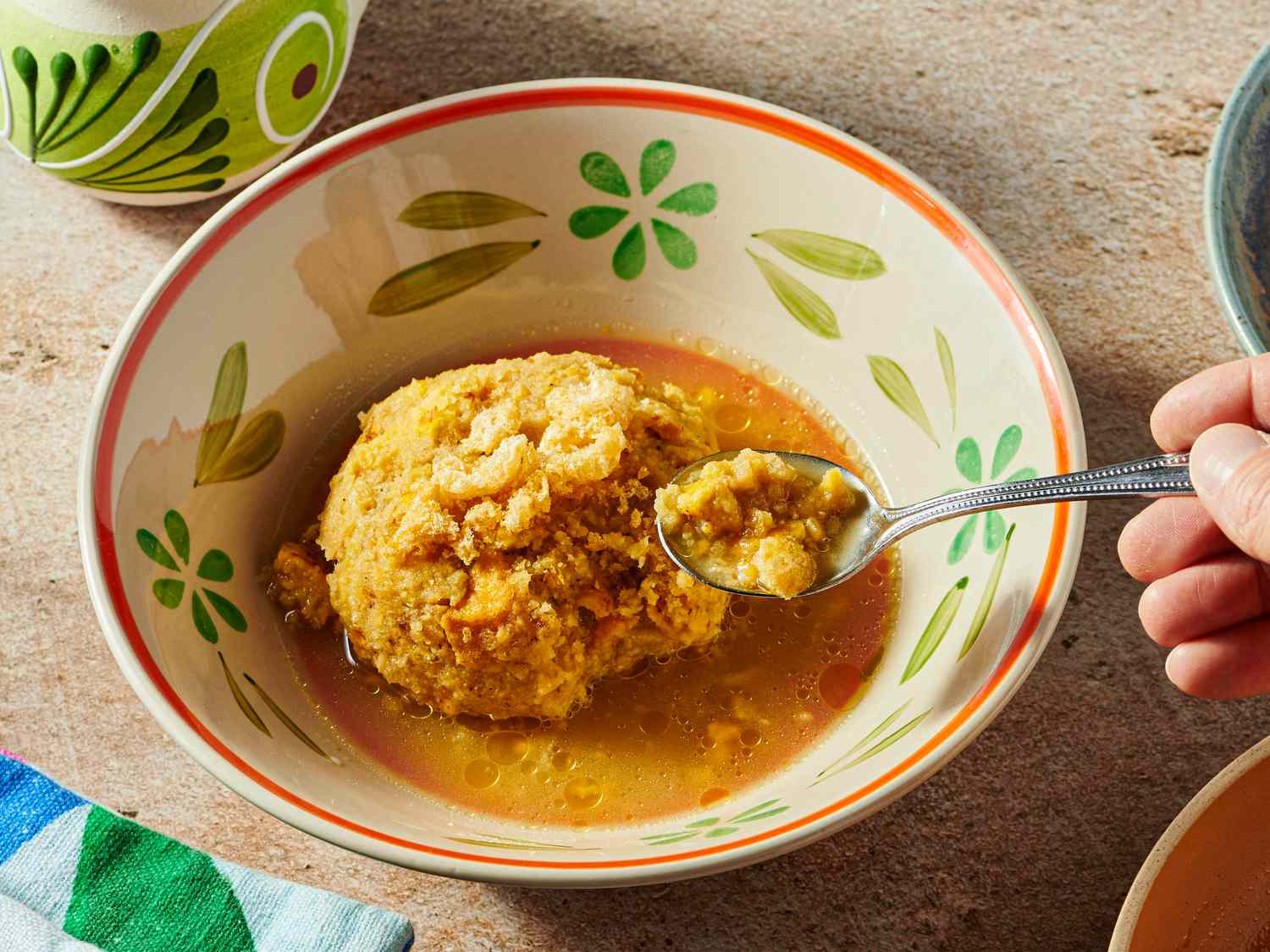 Bowl of Mofongo in a bone colored dish with flowers on it, with a hand holding a spoon of mofongo above bowl. Other dishes and textile on the sides of frame. 