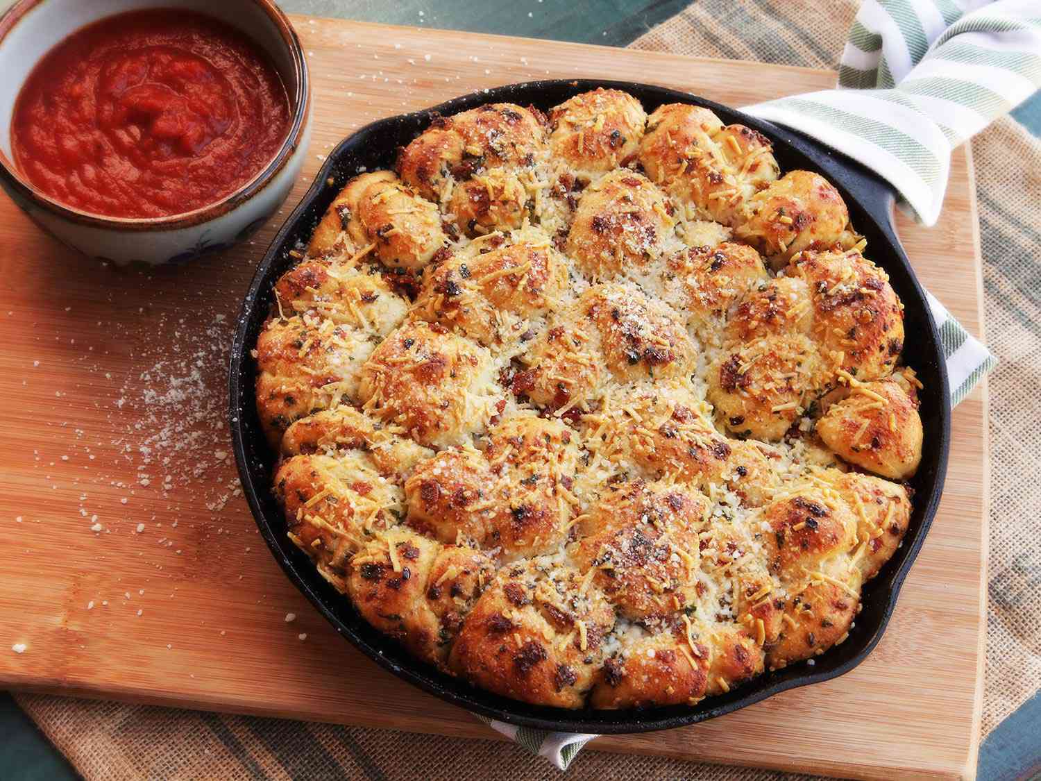A cast iron pan of cheese-covered pepperoni garlic knots with marinara dipping sauce