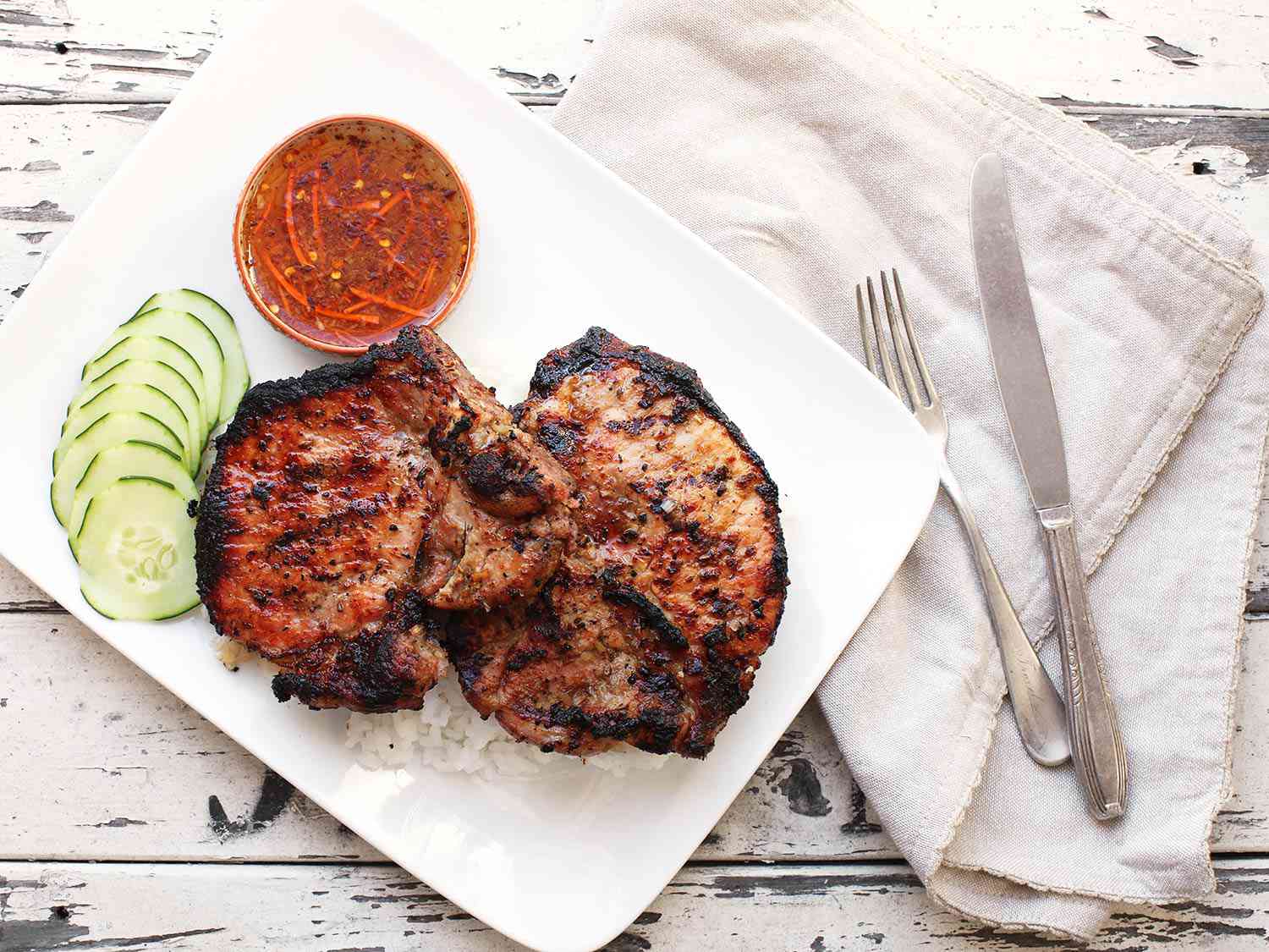 Grilled lemongrass pork chops on a bed of rice on a white plate with sliced cucumbers and nuoc cham on the side.