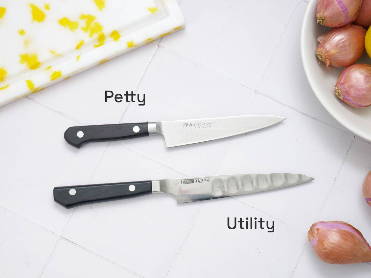 Two kitchen knives labeled as petty and utility placed on a counter near shallots and a cutting board