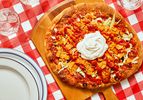 Taco pizza with sour cream on a wooden board, set on a red-checkered tablecloth, plates, and glasses nearby.