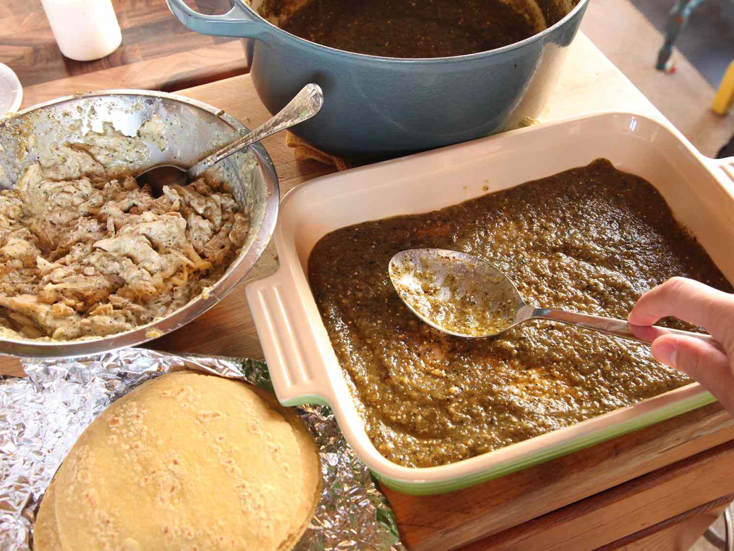 Sauce, chicken filling, and tortillas are arranged next to a baking dish, the bottom of which has been covered in a thin layer of sauce.