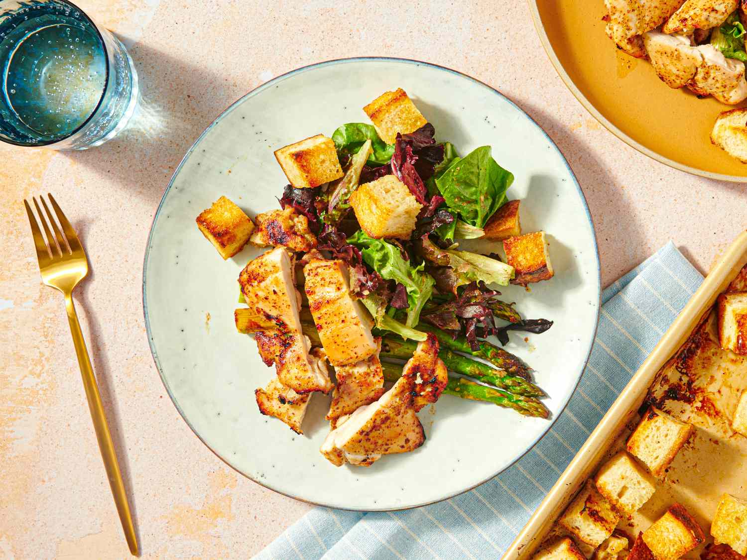 A plate with honey mustard chicken thighs roasted vegetables and fresh greens next to a glass of water and a gold fork