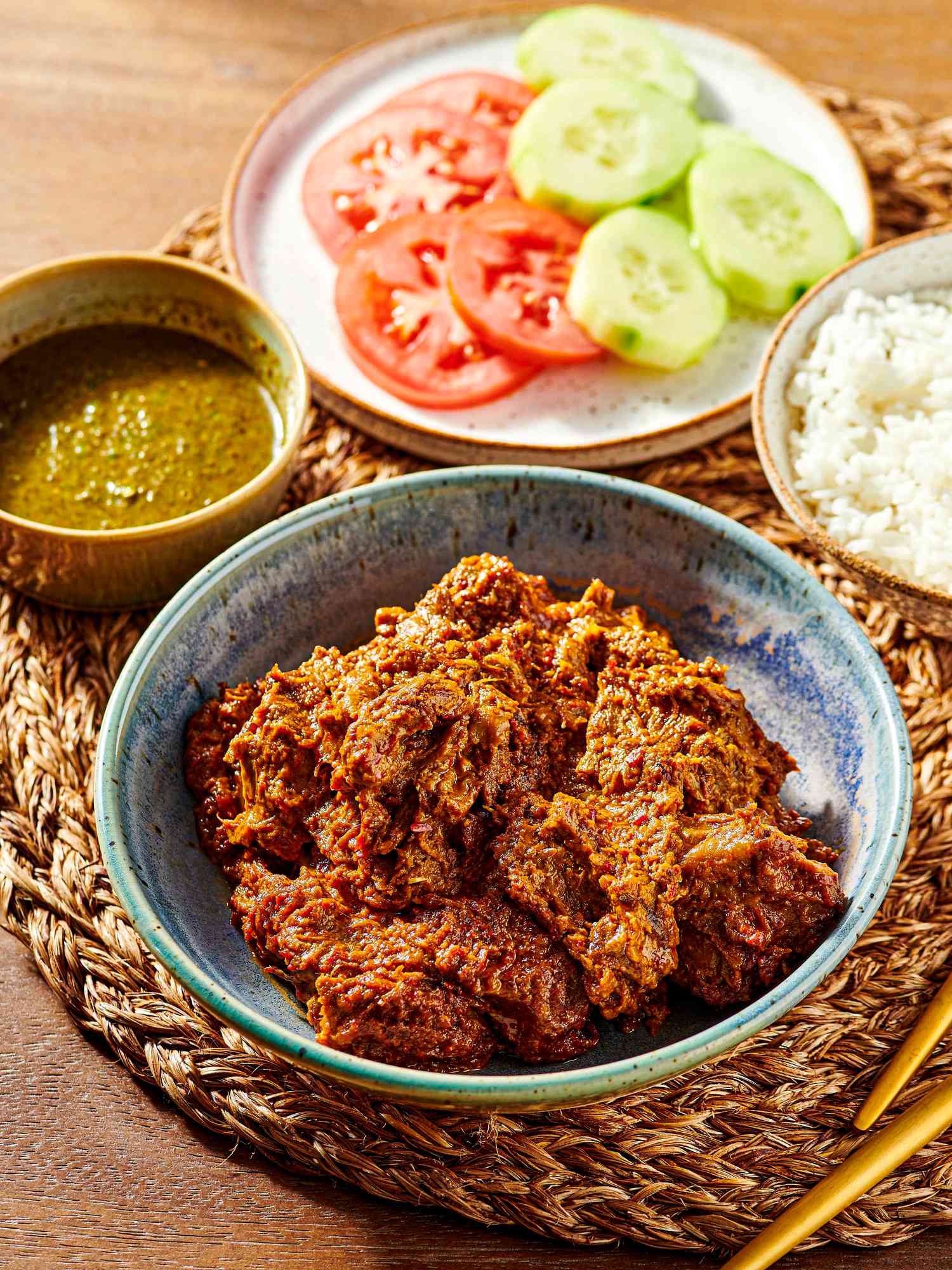 Plate of beef Rendang with rice, cucumbers, tomatoes and crisps on other plates, with a woven placemat and gold cutlery