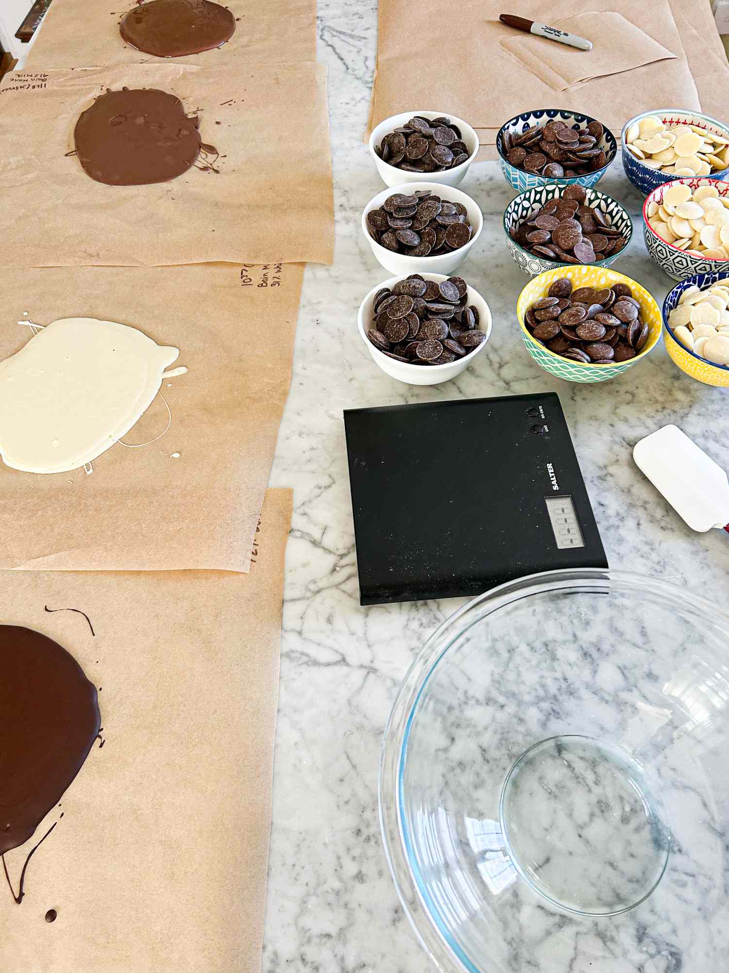 Table with melted chocolate on parchment paper bowls of chocolate pieces a digital scale and a glass bowl