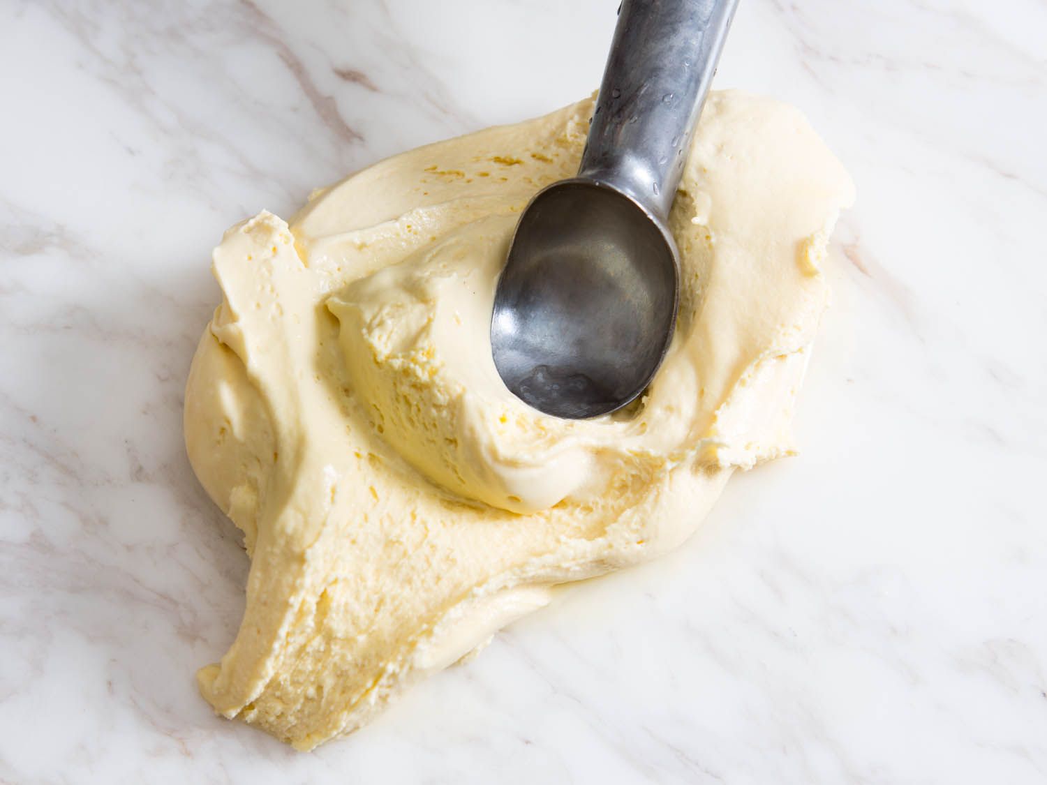 An ice cream scoop pressing on a slab of chewy New England-style ice cream on a marble surface. 