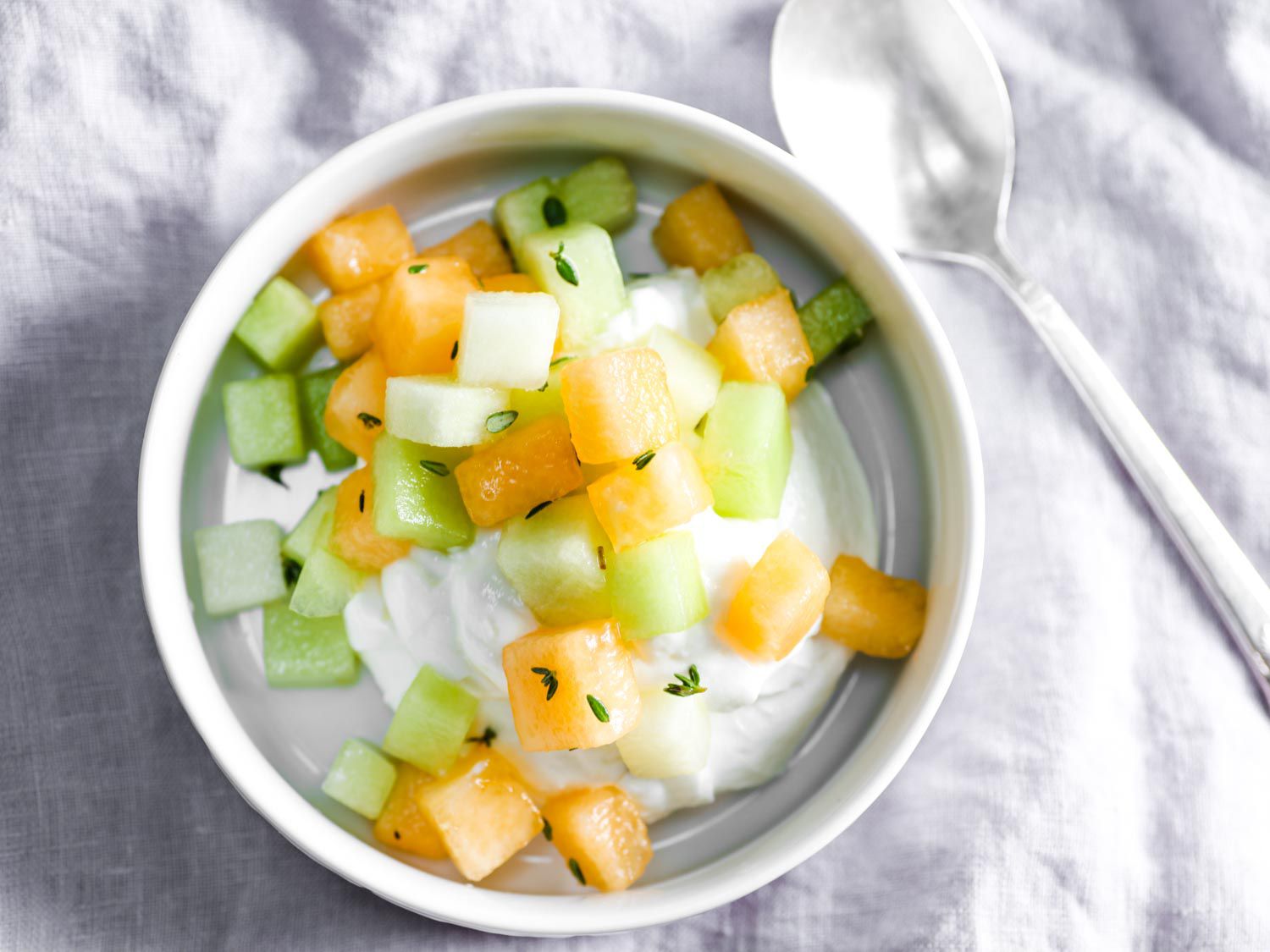 Overhead view of a white bowl containing fresh cantaloupe and honeydew salad with lemon and thyme, served over a scoop of high-quality ricotta.