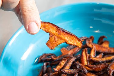 A person holds a piece of cooked mushroom over a blue bowl containing more cooked mushroom pieces