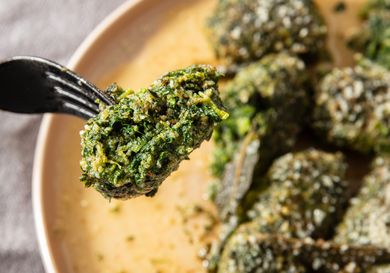 A fork holding a piece of cooked spinach dumpling with more dumplings on a plate in the background