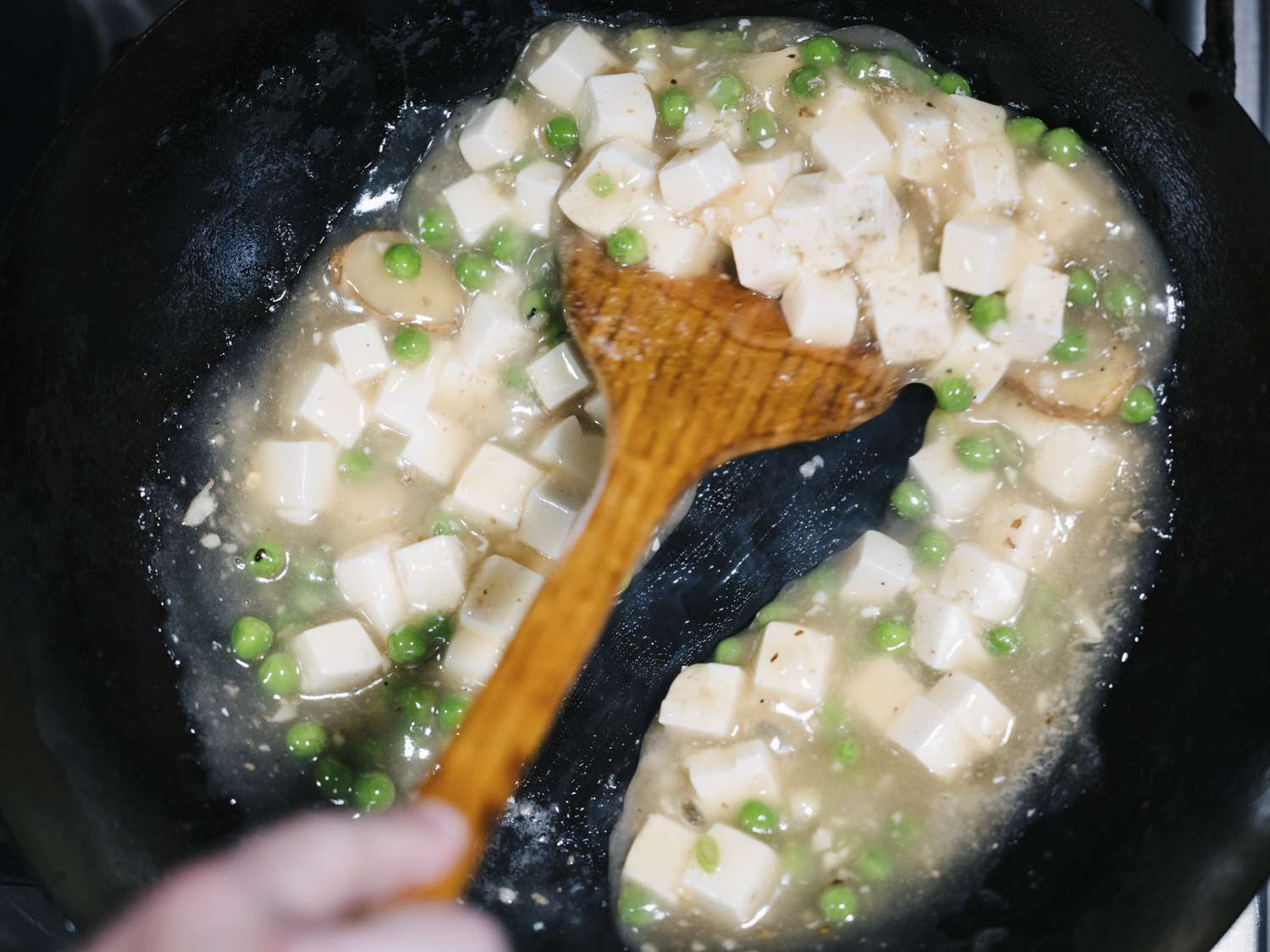 A spatula stirring peas, tofu, and cornstarch slurry together in a wok, showing the thickened consistency of the sauce.