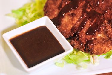 A small white square ramekin of tonkatsu sauce next to a fried cutlet. 