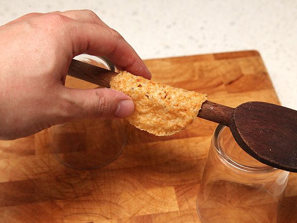 A hand shaping crispy melted cheese disc over the handle of a wooden spoon propped between two inverted glasses to form a taco shell.