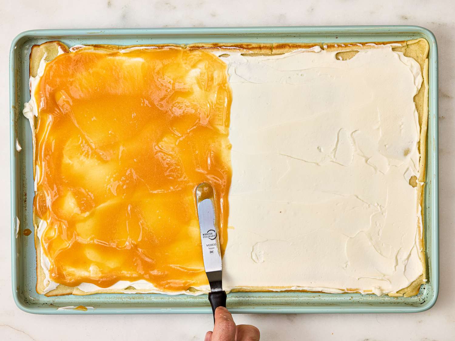 A tray with a halfspread layer of peach topping and cream filling being smoothed with an offset spatula