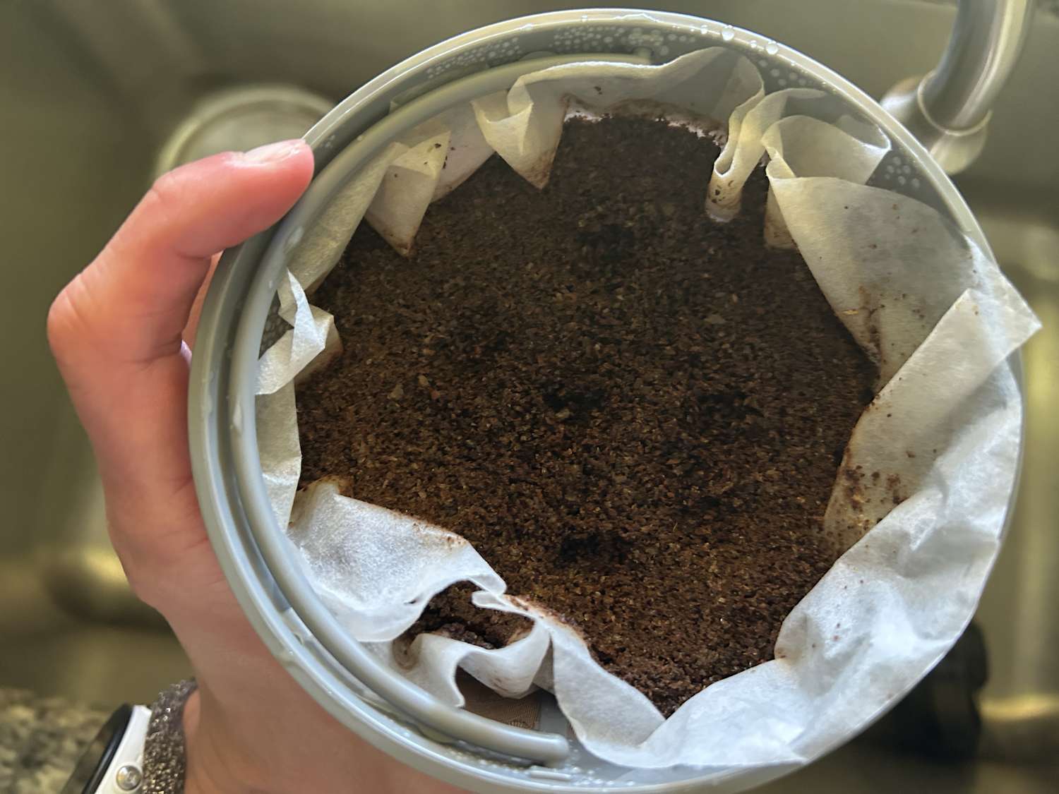 A person holding a cup of coffee grounds in a paper filter over a sink.