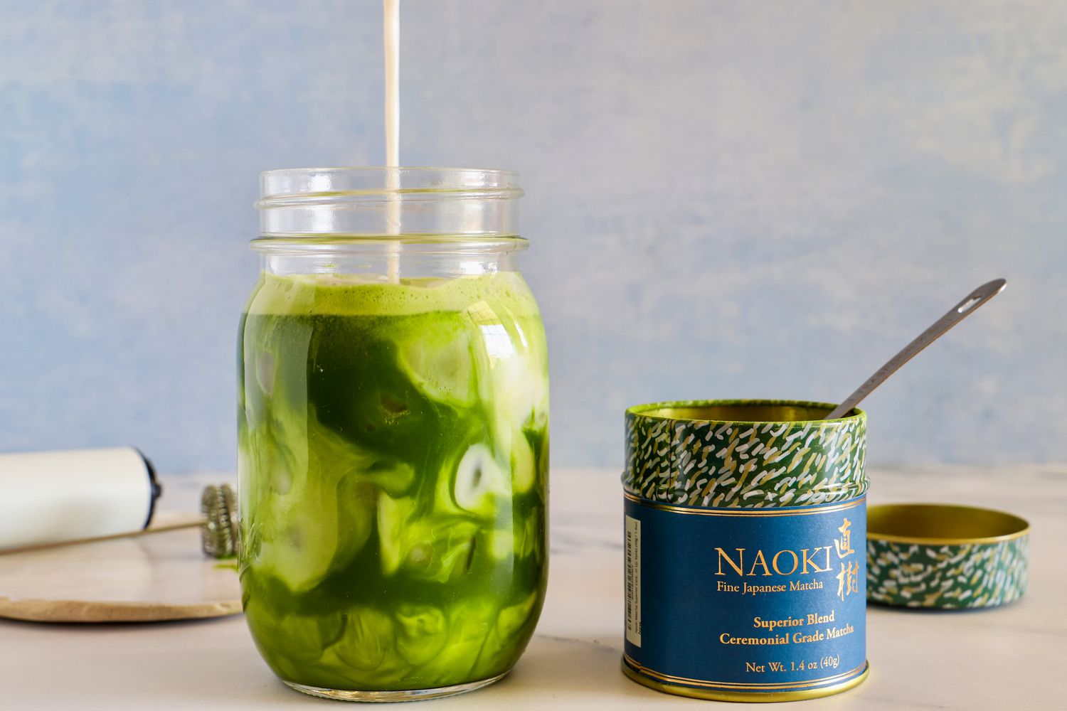 Milk being poured into a glass of Naoki Matcha Superior Ceremonial Blend, next to the container