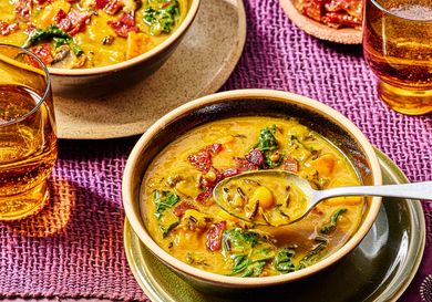 Bowls of pumpkin soup with wild rice and greens spoons placed in one bowl glasses of drink nearby