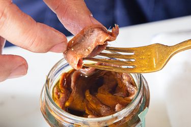 A hand holding anchovies from a jar with a fork