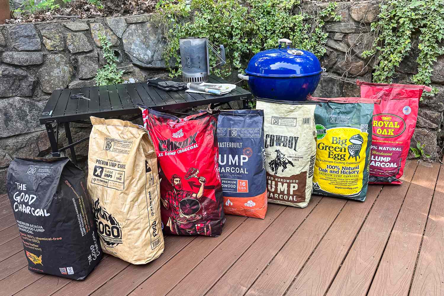 Seven bags of lump charcoal lined up in front of a kettle grill on a deck.
