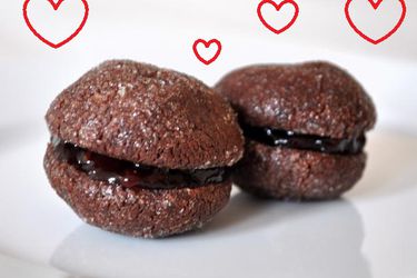 Closeup side view of two chocolate raspberry baci di dama cookies. 4 red, heart-shaped silhouettes have been added to the image.