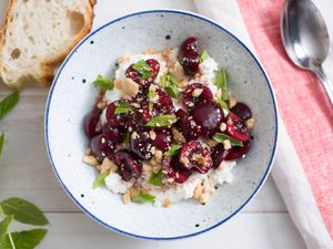 Overhead view of sweet-sour macerated cherries with Marcona almonds, mint, and ricotta, served in as white bowl with blue specks..
