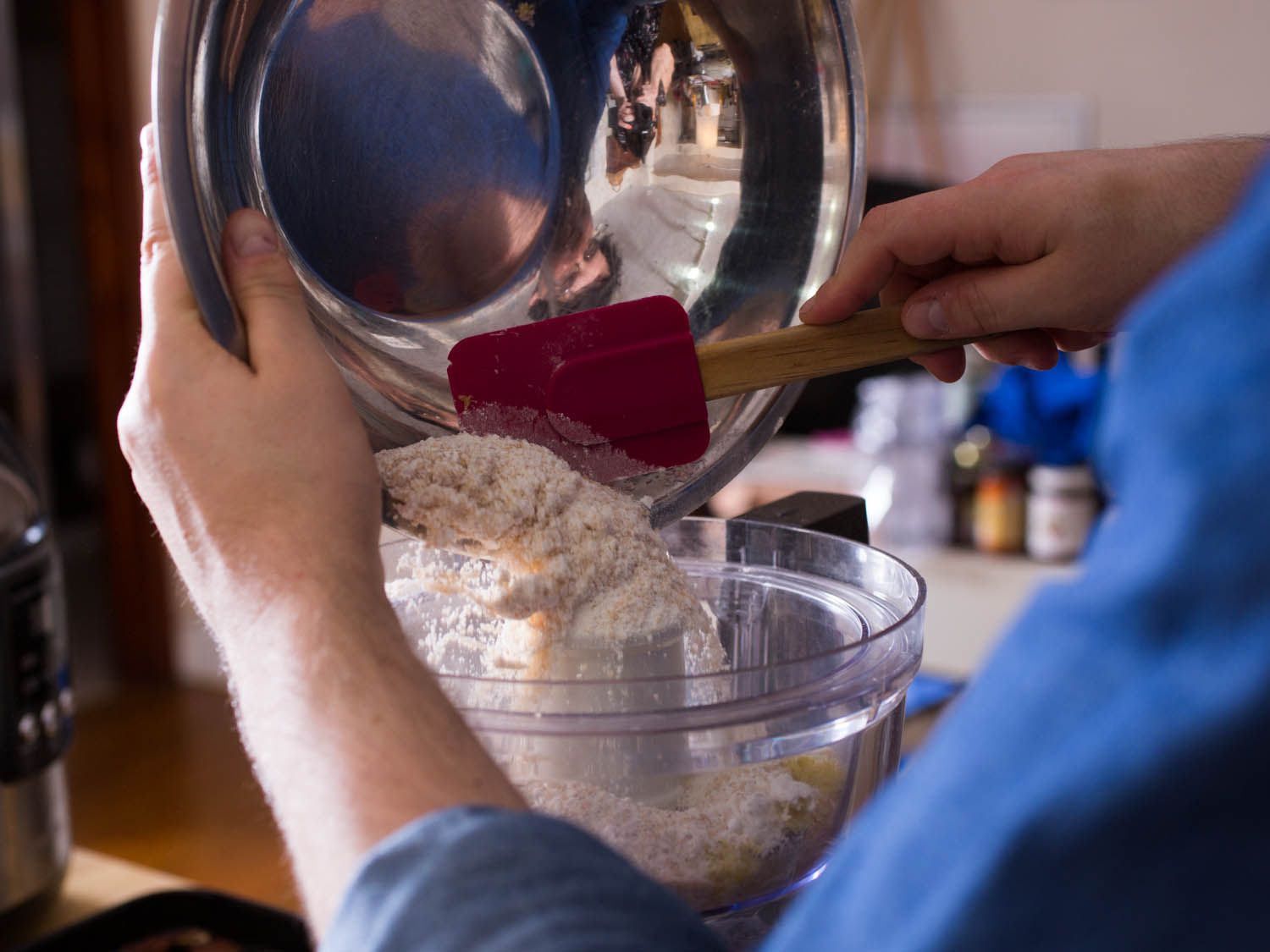 Using a flexible spatula to scrape a mixture of flour, sugar, and lemon zest from a metal bowl into the bowl of a food processor.