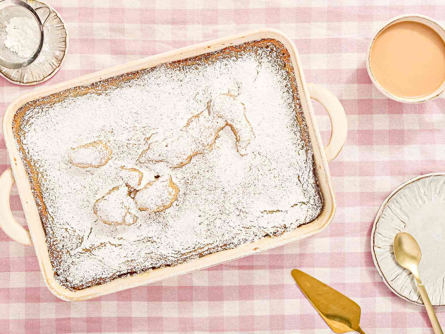 A gooey butter cake in a rectangular dish with powdered sugar topping on a pink gingham tablecloth, surrounded by serving utensils and plates