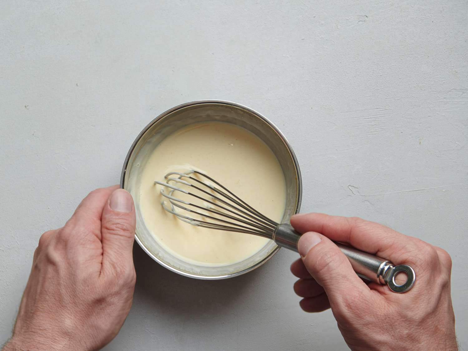 Two hands whisking together the ingredients which form the dressing for the potato salad. The right hand is holding the whisk, which is placed into the bowl of combined ingredients.