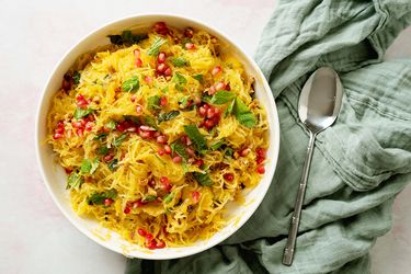 Spaghetti sqwuash with pomegranate, toasted hazelnuts, and mint in a serving bowl with a spoon set on a kitchen towel alongside
