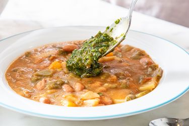 A bowl of vegetable soup being garnished with a green herb mixture a spoon adding the topping
