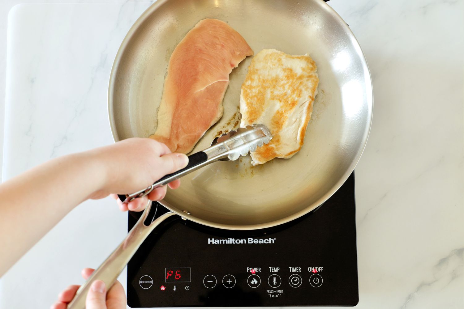 An person flipping a chicken breast cooking in a stainless steel skillet on an induction burner.